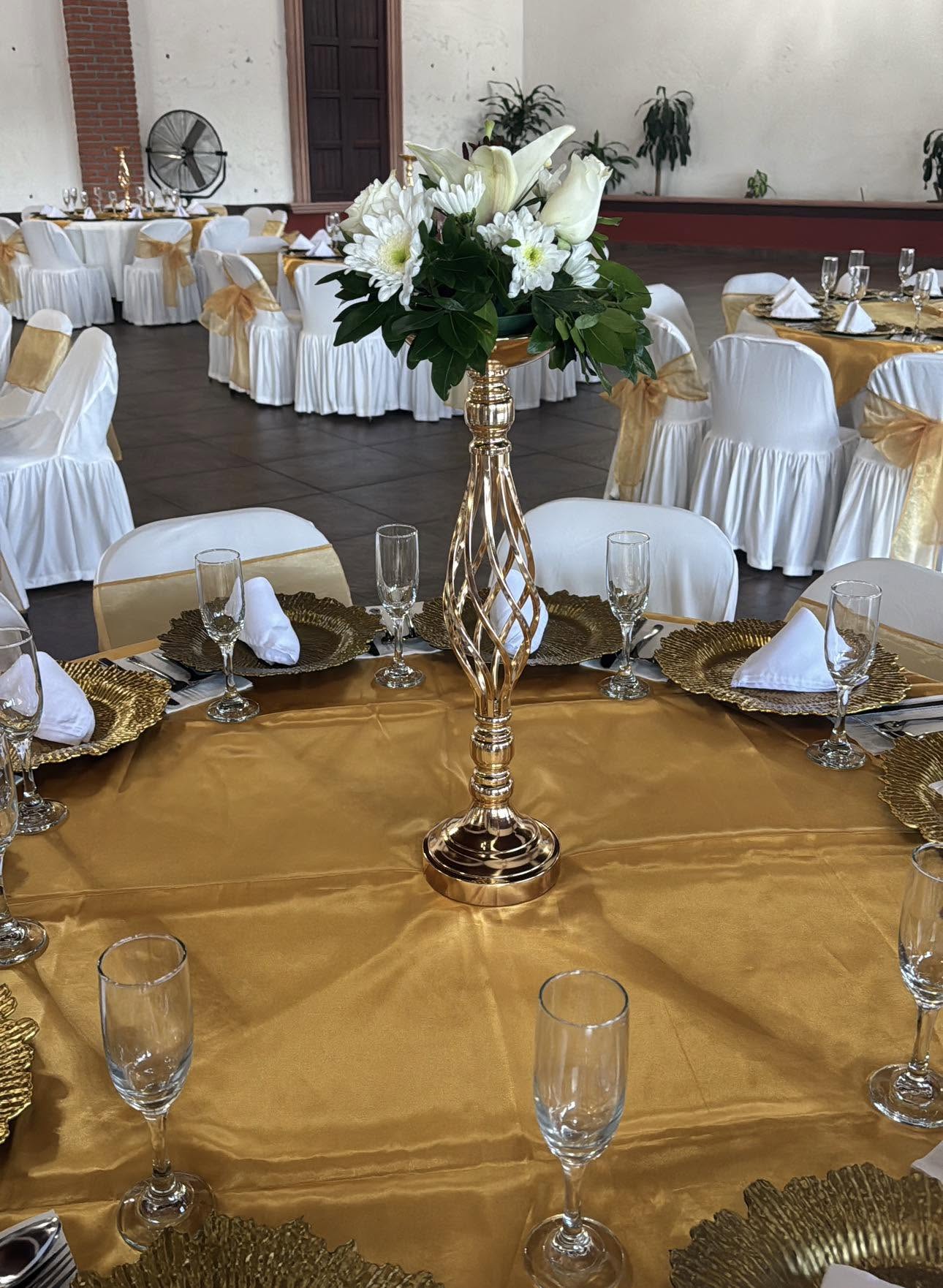 Mesa redonda con mantel dorado, platos base dorados, copas de cristal y centro de mesa con flores blancas